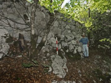 Mike Galaty wearing a blue shirt and khaki pants standing in front of a stone wall with a red and white height marking poll.